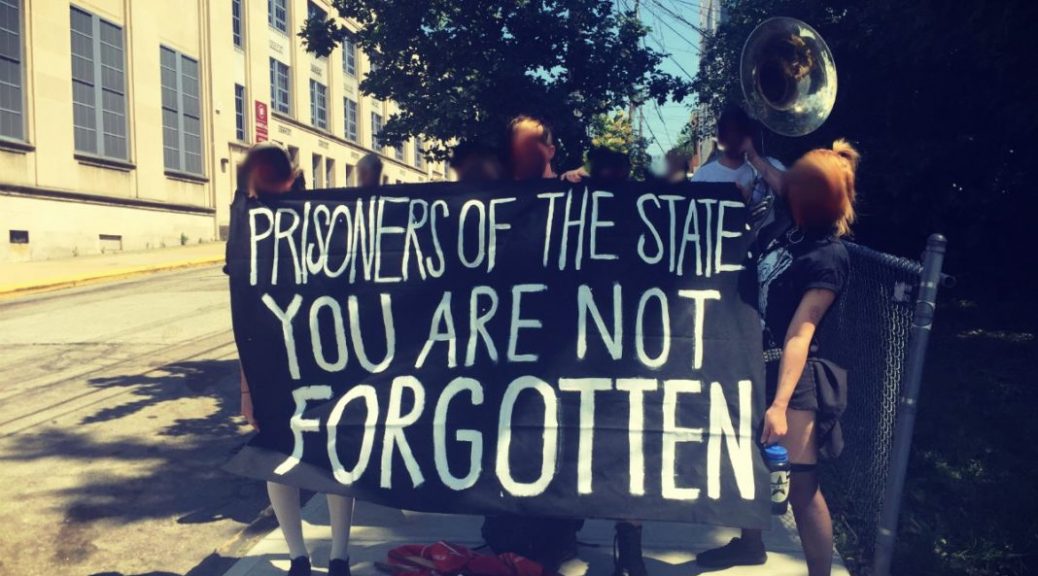 An anti-prison protest in Pittsburgh with people holding musical instruments and a banner reading "Prisoners of the state: you are not forgotten"