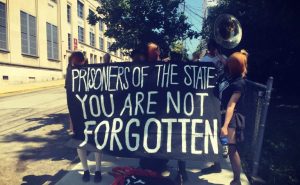 An anti-prison protest in Pittsburgh with people holding musical instruments and a banner reading "Prisoners of the state: you are not forgotten"