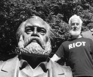 Joshua Clover with a beard and shirt reading "Riot" next to a statue of Karl Marx