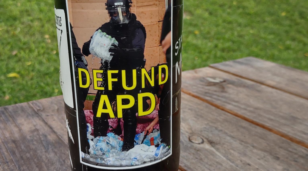 "Defund APD" sticker on a water bottle, depicting an asheville police officer stabbing and crushing water bottles after raiding a medic table during George Floyd protests in 2020