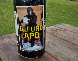 "Defund APD" sticker on a water bottle, depicting an asheville police officer stabbing and crushing water bottles after raiding a medic table during George Floyd protests in 2020. Based on a photo by Angie Wilhelm