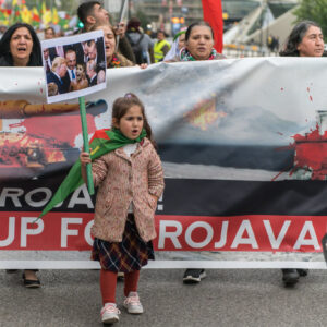Mostly women carrying "Stand Up For Rojava" banner with a small girl and a sign picturing world leaders leaning in on a small Kurdish child
