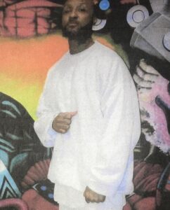A picture of Mwalimu Shakur in white sweat pants and shirt in front of a mural