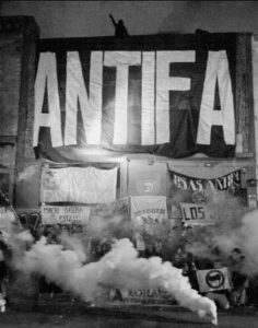 a black and white shot from a demonstration with smoke bombs, flares and large banner reading "Antifa" in front of La Cultura Del Barrio social space & gym