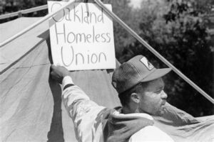 black and white photo of someone putting up a sign reading "Oakland Homeless Union" in front of a tent